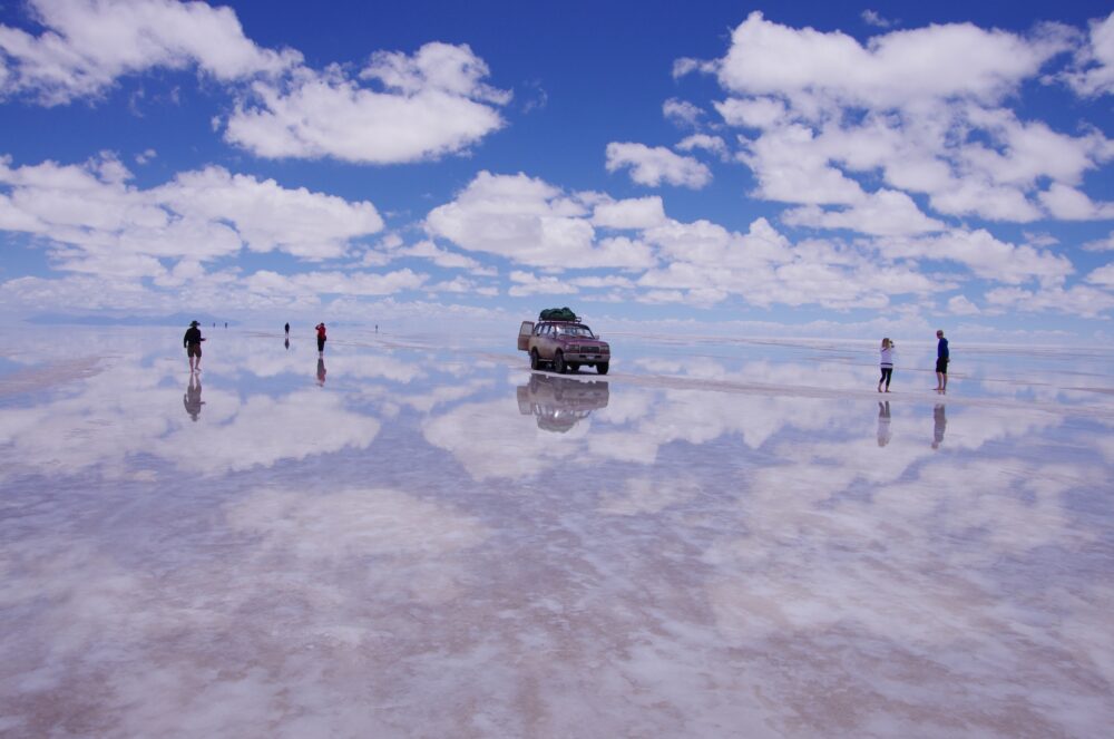 ボリビア・ウユニ塩湖で雲を映す鏡張りの大地と四輪駆動車、観光客が歩く絶景風景