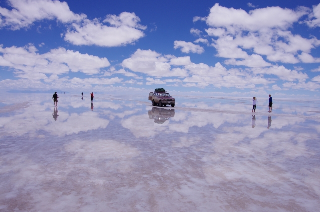 ウユニ塩湖の雨季に現れる鏡張りの風景。空と雲が水面に反射し、人物や車が上下対称に映っている様子