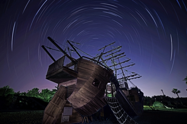 星の軌跡が描かれる夜空の下にある木造帆船。位置を測れなかった時代の航海を象徴する光景。