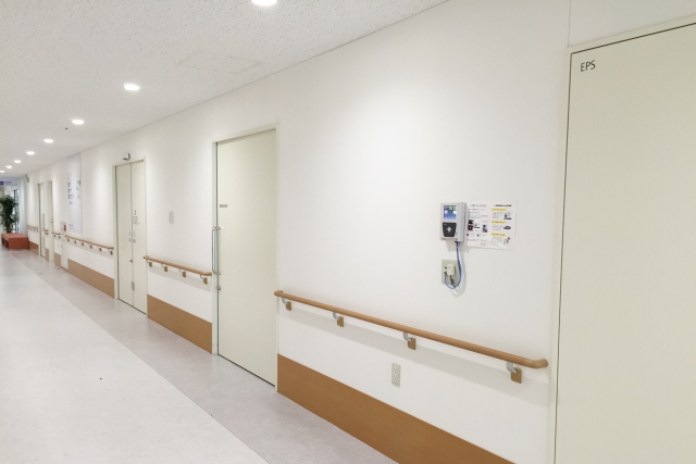 白い壁と複数の扉が並ぶ病院の通路。診察室や検査室が並び、行き先に迷いやすい院内廊下